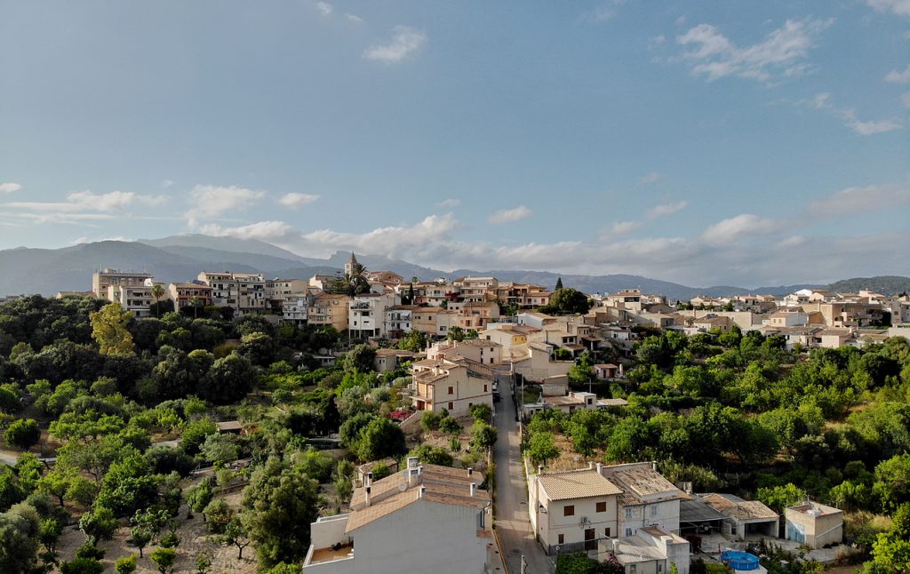 Panoramablick auf Campanet mit sandfarbenen Häusern, Kirchturm, viel Grün und den Bergen der Tramuntana unter blauem Himmel.