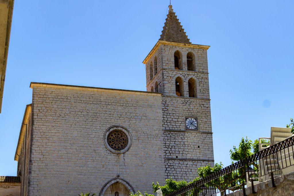 Steinernes Kirchenschiff mit Rosettenfenster und quadratischem Glockenturm mit Uhr in Campanet vor blauem Himmel.