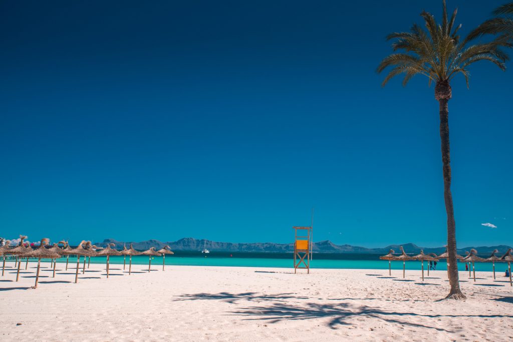 Breiter Strand mit Strohdachschirmen, gelbem Rettungsturm und türkisblauem Meer vor Bergkulisse in Can Picafort.