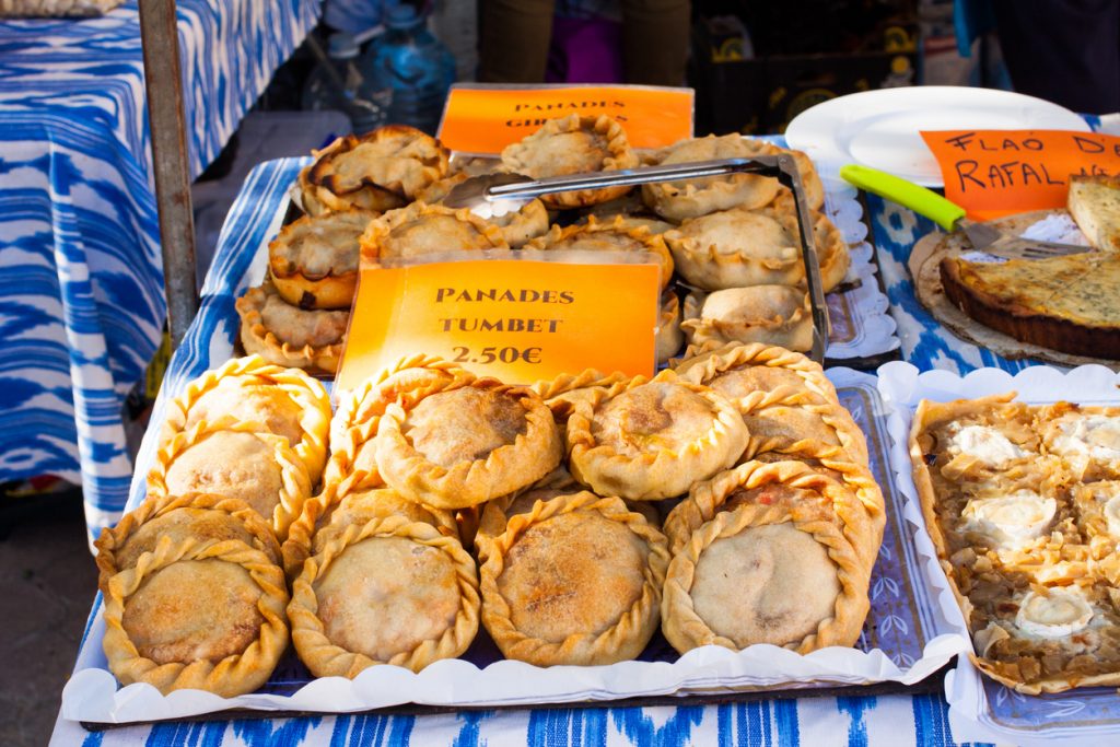Stapel runder, gefüllter Panades mit geflochtenem Rand auf Tabletts; Schild „Panades Tumbet 2.50 €“, daneben Zange und weiteres Gebäck auf einem Marktstand.