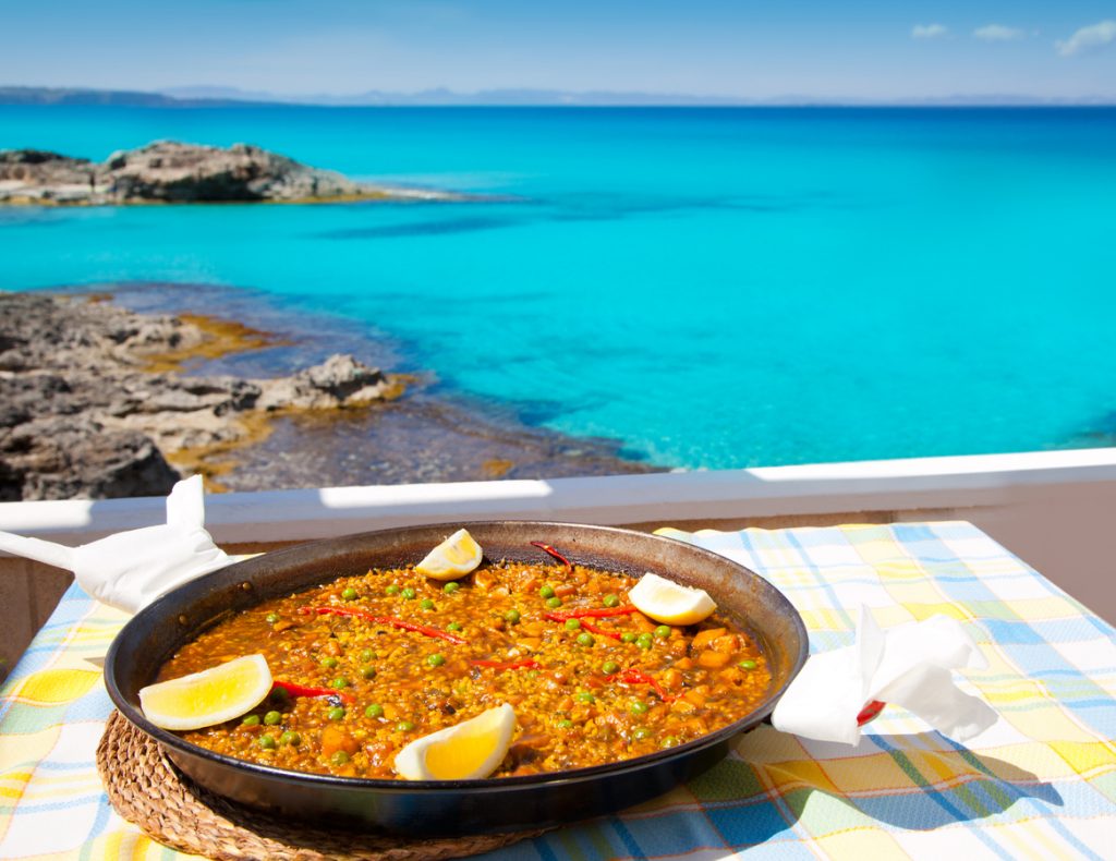 Paellapfanne mit Zitronenspalten auf karierter Tischdecke vor türkisblauem Meer und Felsen auf Formentera.