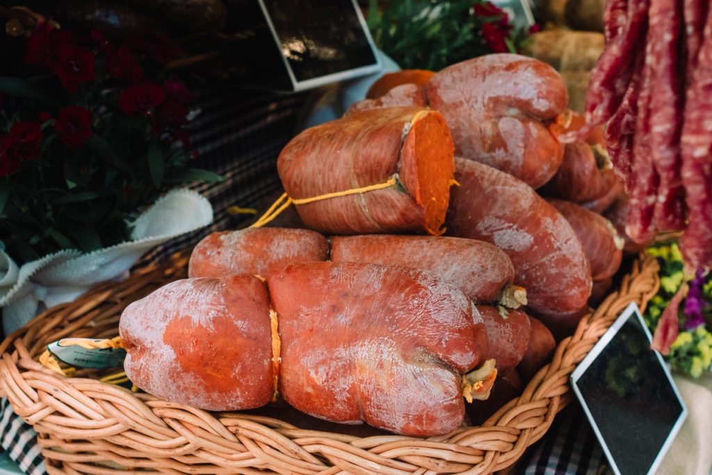 Mehrere grobe Sobrassada-Würste in einem geflochtenen Korb; eine angeschnitten mit kräftig rot-orangefarbener Füllung, die Naturhülle mehlig bestäubt, daneben weitere Wurstwaren und Deko am Marktstand.