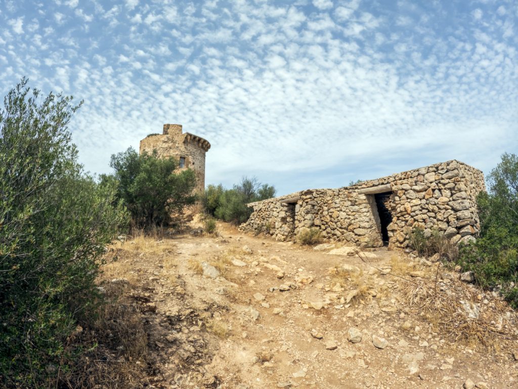 Steinerner Wachturm mit Trockensteinhütte auf Mallorca, umgeben von Macchia und Felsen.