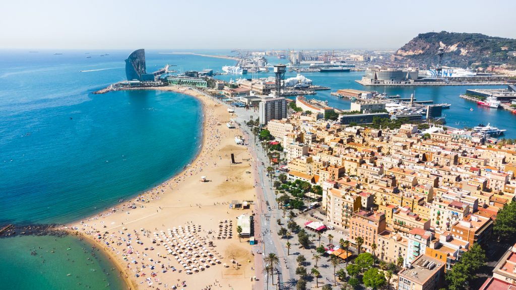 Luftaufnahme von Barceloneta mit goldenem Sandstrand, W-Hotel und dem Hafen von Barcelona bei klarem Himmel.
