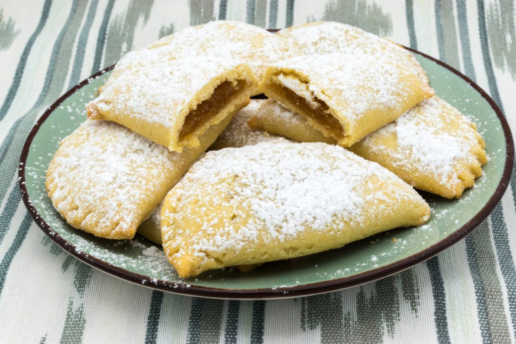 Mehrere mit Puderzucker bestäubte Robiols liegen auf einem grünen Teller, teils halb geöffnet mit sichtbarer goldener Füllung.