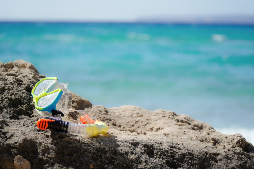 Eine Schnorchelmaske mit Schnorchel liegt auf einem Felsen am Strand von Formentera vor türkisfarbenem Wasser.