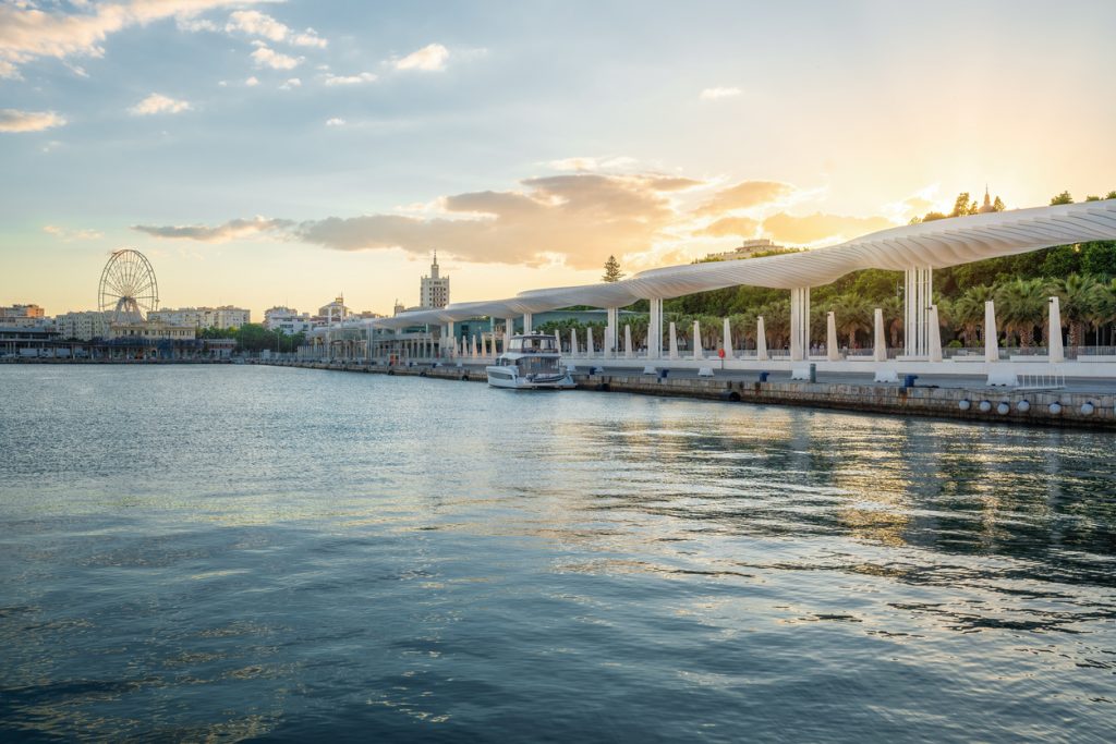 Wellenförmige weiße Pergola am Muelle Uno mit Promenade, Boot und Stadt im Abendlicht.