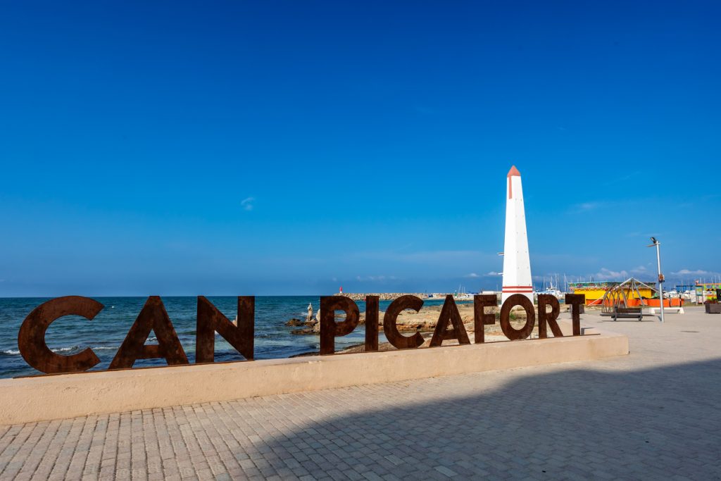 Großer Metall-Schriftzug „CAN PICAFORT“ an der Promenade mit weiß-rotem Obelisken, Meer und Hafen im Hintergrund.