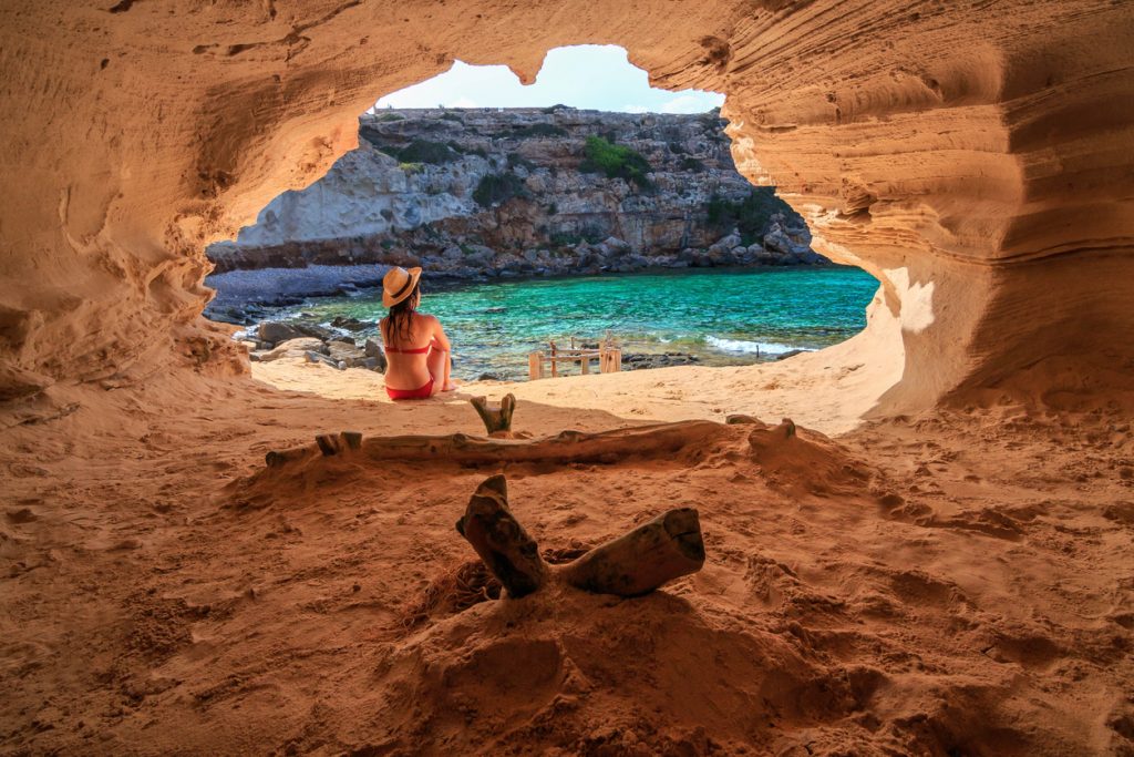 Aus einer sandigen Felsgrotte blickt eine Person mit Hut auf die türkisblaue Cala en Baster mit Felswänden.