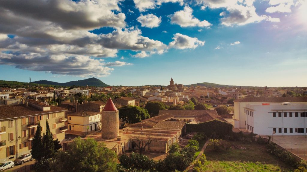 Luftaufnahme von Algaida auf Mallorca mit runder Windmühle mit rotem Dach, steinernen Häusern und Hügeln unter blauem Himmel.