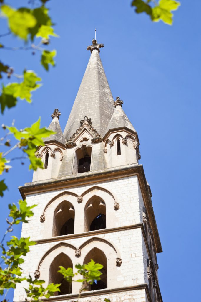 Nahaufnahme des steinernen Kirchturms in Binissalem mit Spitzdach, Spitzbogenfenstern und grünen Blättern im Vordergrund.