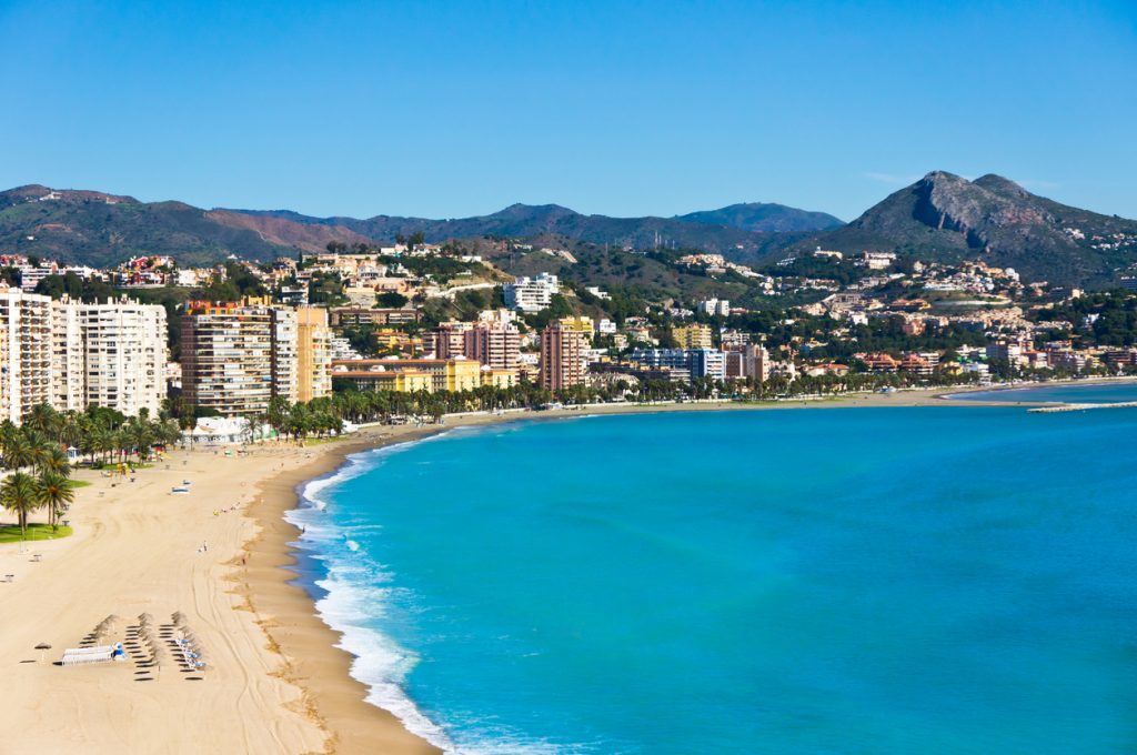 Breiter Sandstrand mit türkisblauem Meer, Promenade und Wohnhäusern vor Bergen in Málaga.