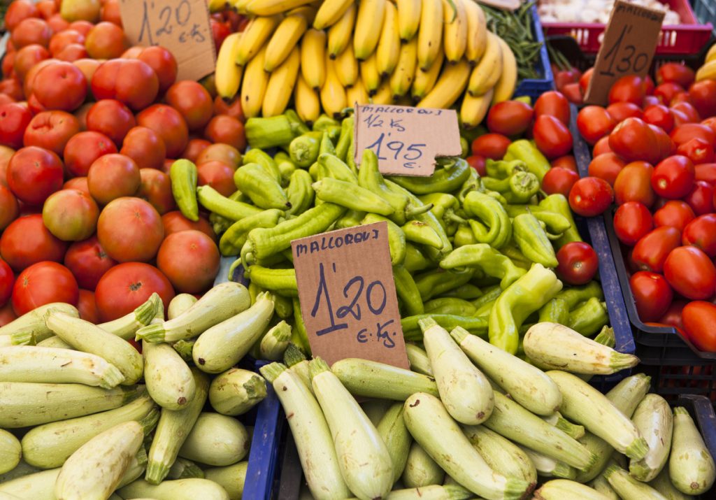 Kisten voller Tomaten, hellgrüner Zucchini, grüner Paprika und Bananen mit handgeschriebenen Euro-Preisschildern auf einem mallorquinischen Markt.