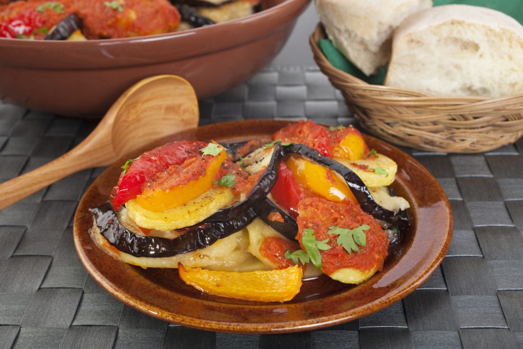 Teller mit Tumbet aus Auberginen, Kartoffeln und Paprika in Tomatensoße, daneben Brotkorb und Holzlöffel.