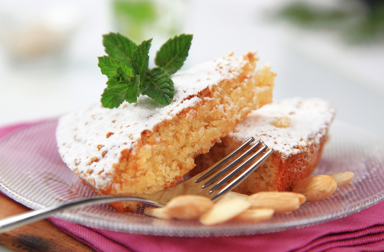 Stück Mandelkuchen mit Puderzucker, Minzzweig und Gabel auf Glasteller.