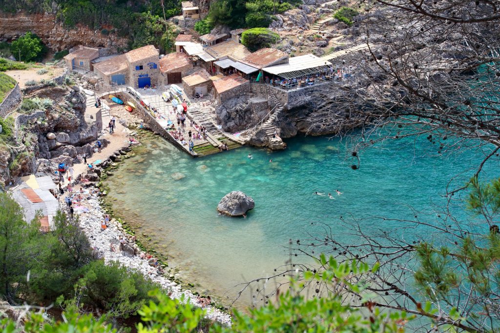 Türkise Cala Deià mit Badenden und Steinhäusern am Ufer.