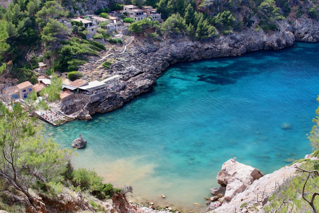 Cala Deià mit türkisblauem Wasser, Felsen und Steinhäusern.