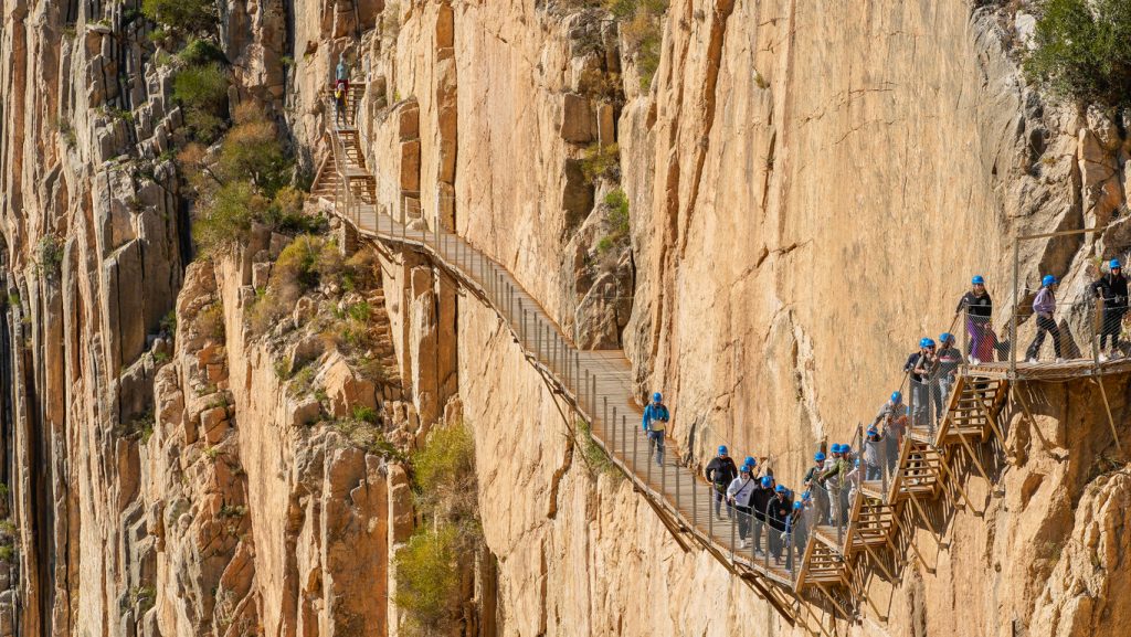 Wanderer mit Helmen gehen über den schmalen Holzsteg an der steilen Felswand.