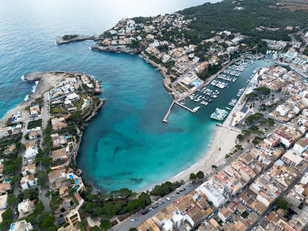 Luftaufnahme der geschützten Bucht von Porto Cristo mit Hafen, Strand und türkisblauem Wasser.