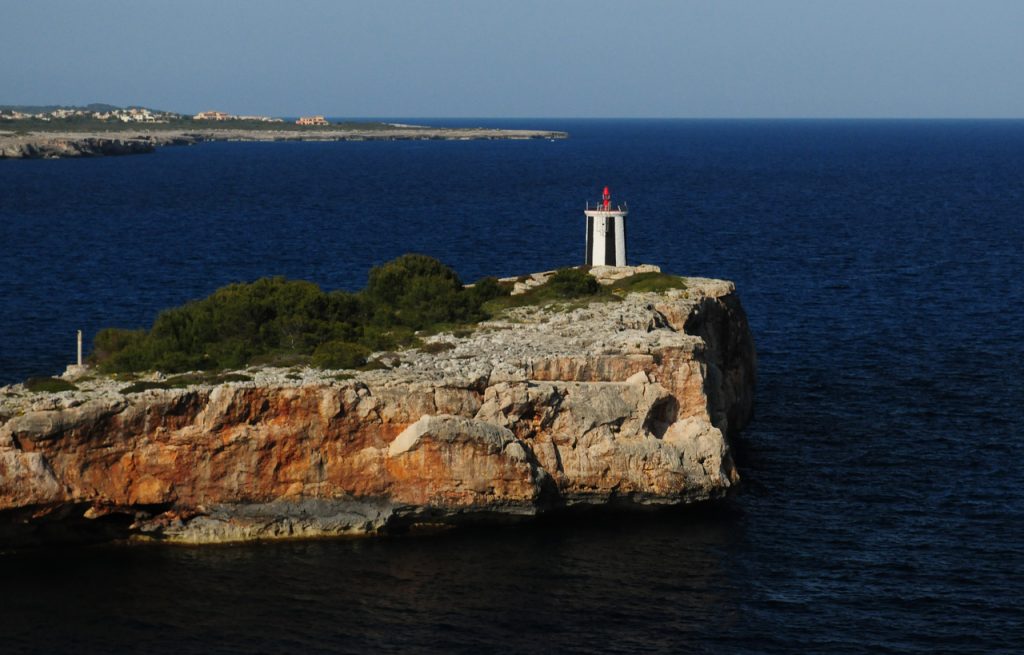 Kleiner weißer Leuchtturm mit roter Spitze steht auf einer kargen Klippe über dem tiefblauen Meer nahe Porto Cristo.