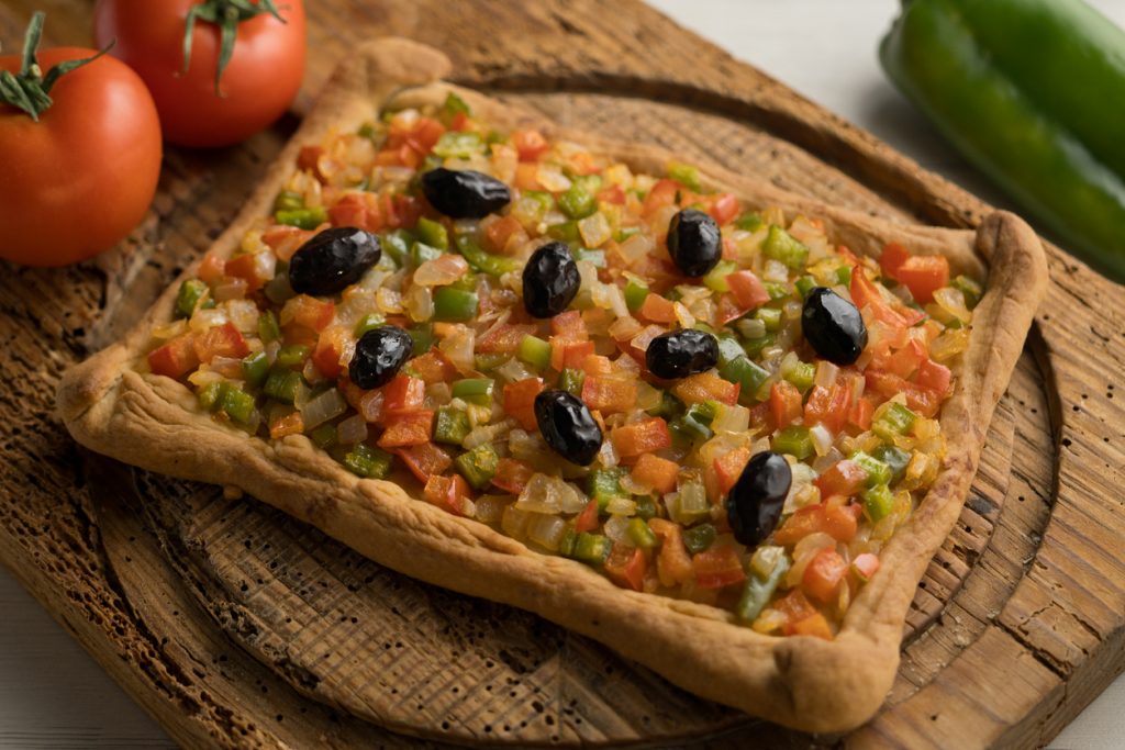 Rechteckige Coca de trampó mit Tomaten-, Paprika- und Zwiebelwürfeln sowie schwarzen Oliven auf einem Holzbrett.