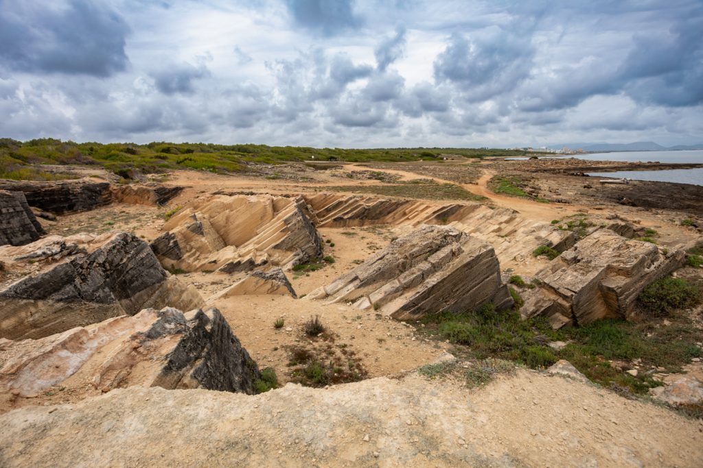 Steinerne Grabkammern und rechteckige Strukturen der Necropolis de Son Real liegen an der rauen Küste unter wolkigem Himmel.