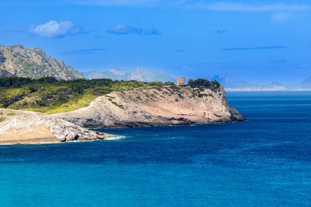 Felsige Landzunge bei Cala Mitjana mit Pinien und altem Wachturm ragt ins türkisblaue Meer, dahinter schimmern Berge.