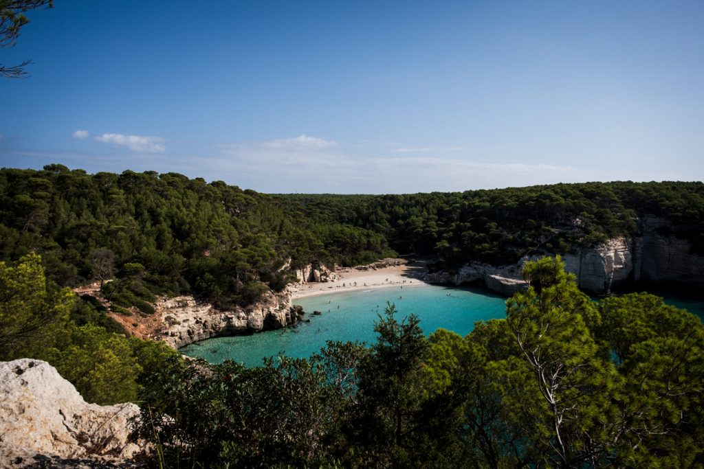 Vom Camí de Cavalls fällt der Blick auf eine geschützte Sandbucht mit weißen Felsen, Pinienwald und türkisblauem Wasser.