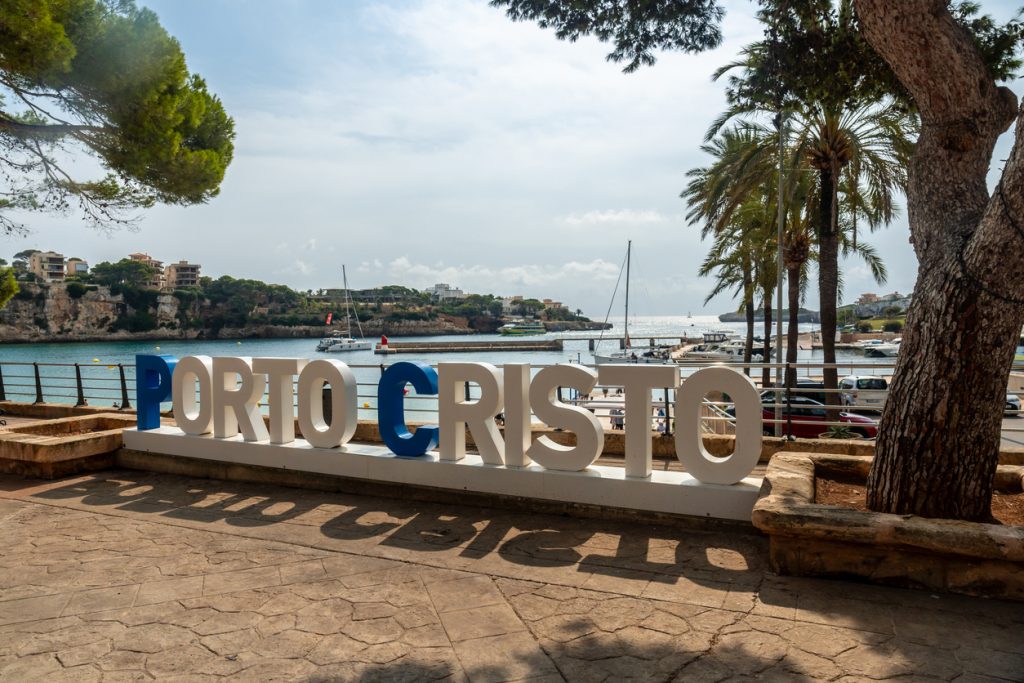 Weiß-blauer „PORTO CRISTO“-Schriftzug vor der Bucht mit Booten, Palmen und Felsenküste.
