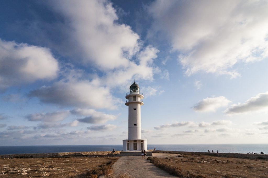 Schlanker weißer Leuchtturm steht auf karger Küste am Cap de Barbaria vor Meer und weitem Himmel.