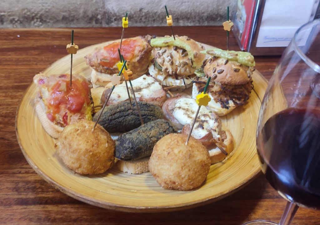 Holzteller mit verschiedenen Tapas und Pintxos, darunter Croquetas und belegte Brote, daneben ein Glas Rioja-Crianza in einer Bar