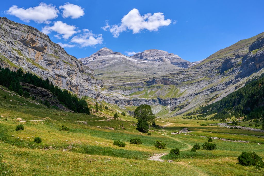 Grüne Talsohle mit Pfad und einzelnen Bäumen liegt zwischen grauen Kalkwänden, dahinter ragen die Gipfel des Monte Perdido unter blauem Himmel.