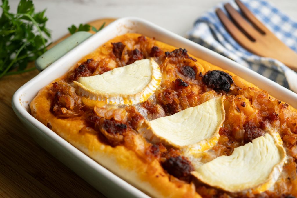 In einer Auflaufform liegt eine goldbraun gebackene Koka mit Sobrasada, geschmolzenem Ziegenkäse und leichtem Honigglanz.