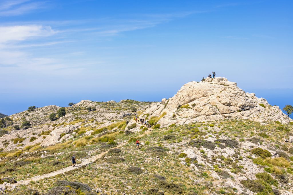 Wanderer ziehen über einen steinigen Höhenpfad zu einer Felskuppel mit Meereshorizont unter blauem Himmel.