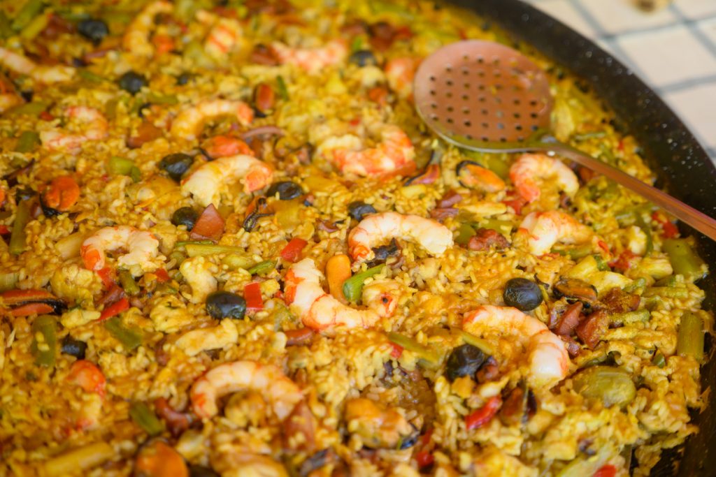 Pfanne mit gelber Paella samt Garnelen, Muscheln, Gemüse und einem Schöpflöffel.