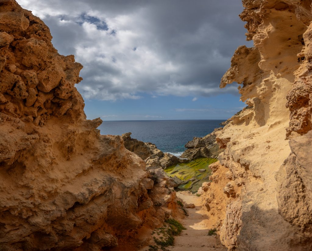 Enge sandfarbene Felsgasse führt zu algenbewachsenen Felsplatten und blauem Meer unter wolkigem Himmel.