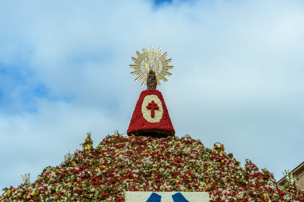 Eine Marienstatue mit goldenem Strahlenkranz steht auf einer riesigen Pyramide aus roten und weißen Blumen.