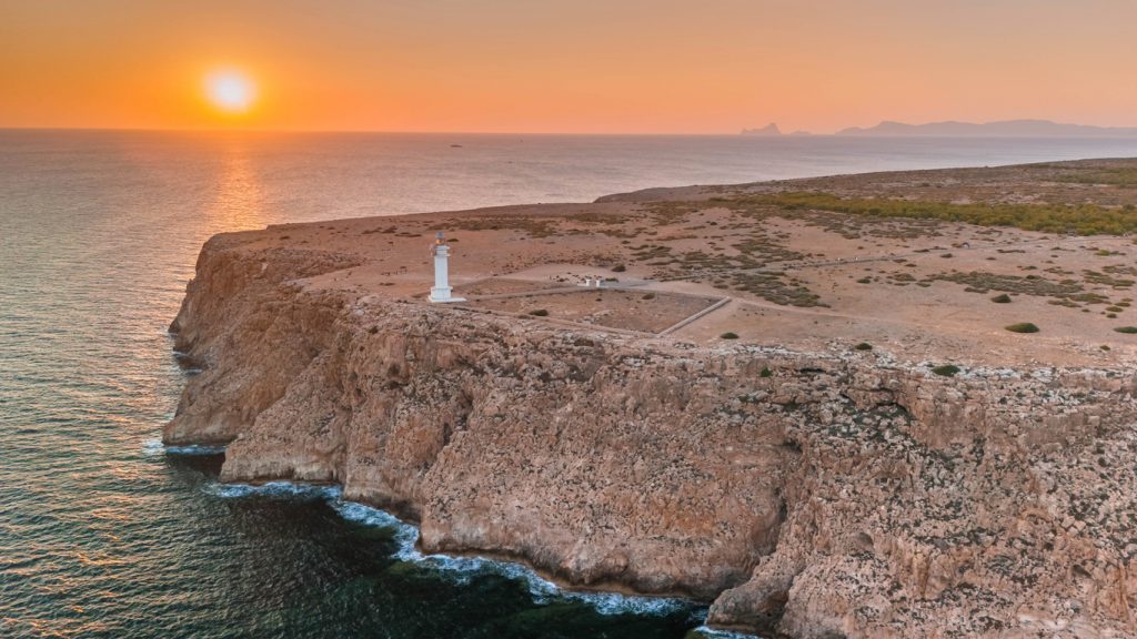Luftaufnahme eines weißen Leuchtturms auf karger Steilküste von Formentera mit untergehender Sonne über dem Meer.