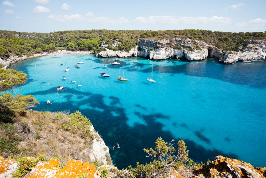 Blick von der Klippe auf Cala Macarella mit Segelbooten im klaren Wasser.
