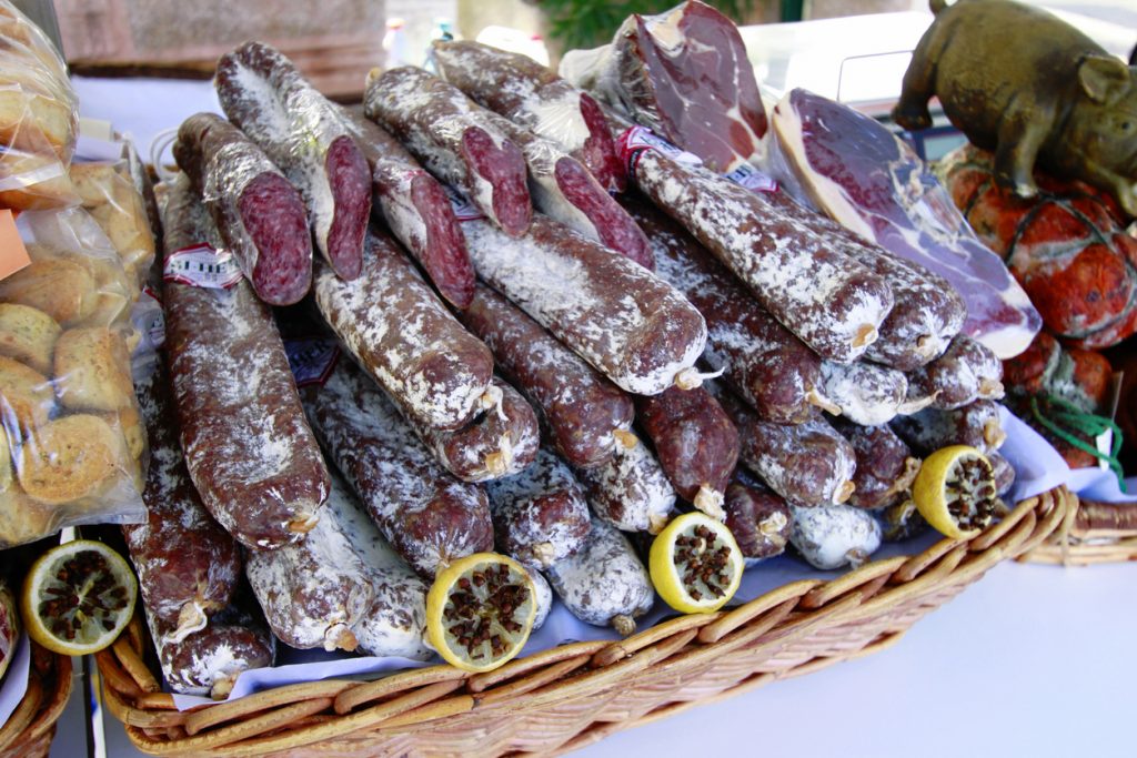 Geflochtener Korb voller weißgereifter, luftgetrockneter Würste mit aufgeschnittenen Enden, flankiert von Schinkenstücken und Zitronen mit Nelken.