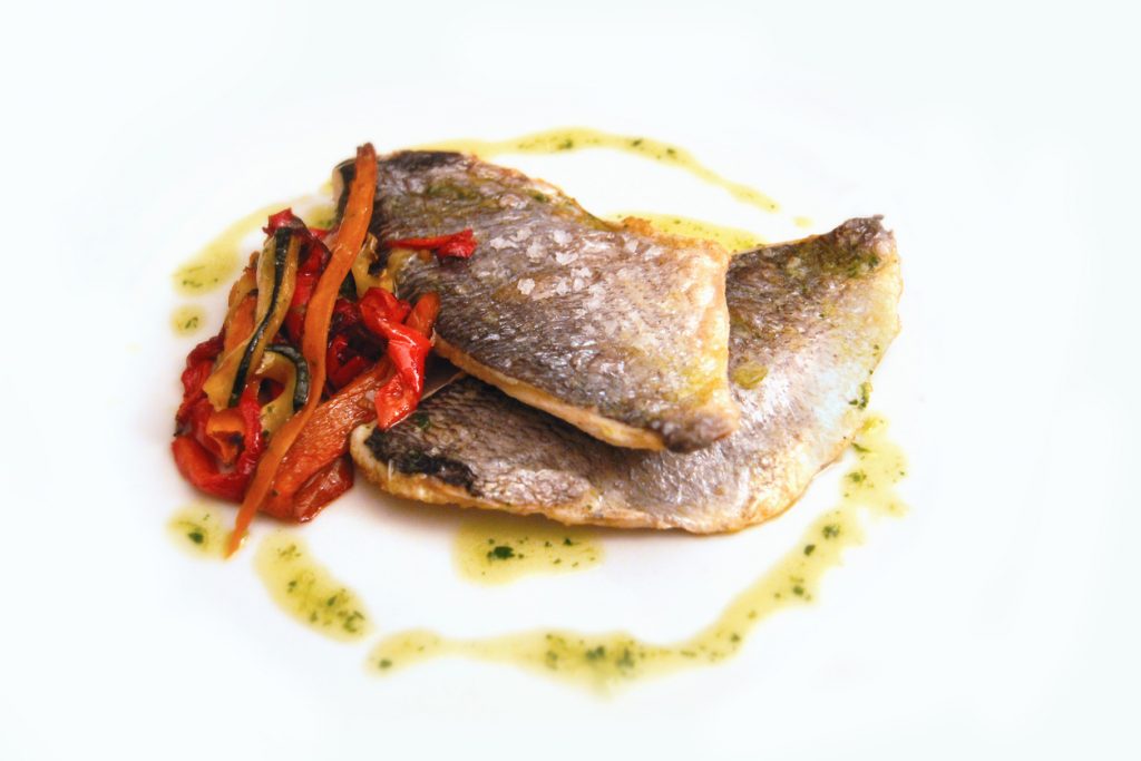 Zwei gebratene Llampuga-Filets mit knuspriger Haut liegen neben geschmorter Paprika, umrahmt von Kräuteröl