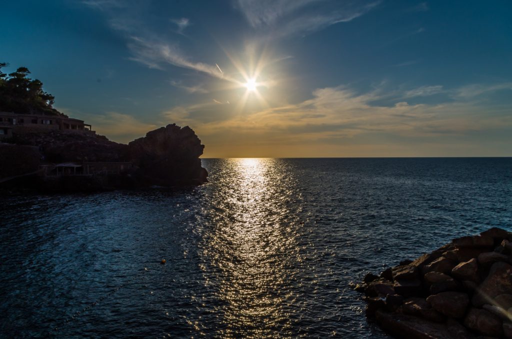 Tiefe Sonne über dem Meer bei Cala Estellencs, goldene Lichtbahn auf dem Wasser, Felsen und Häuser als Silhouetten.