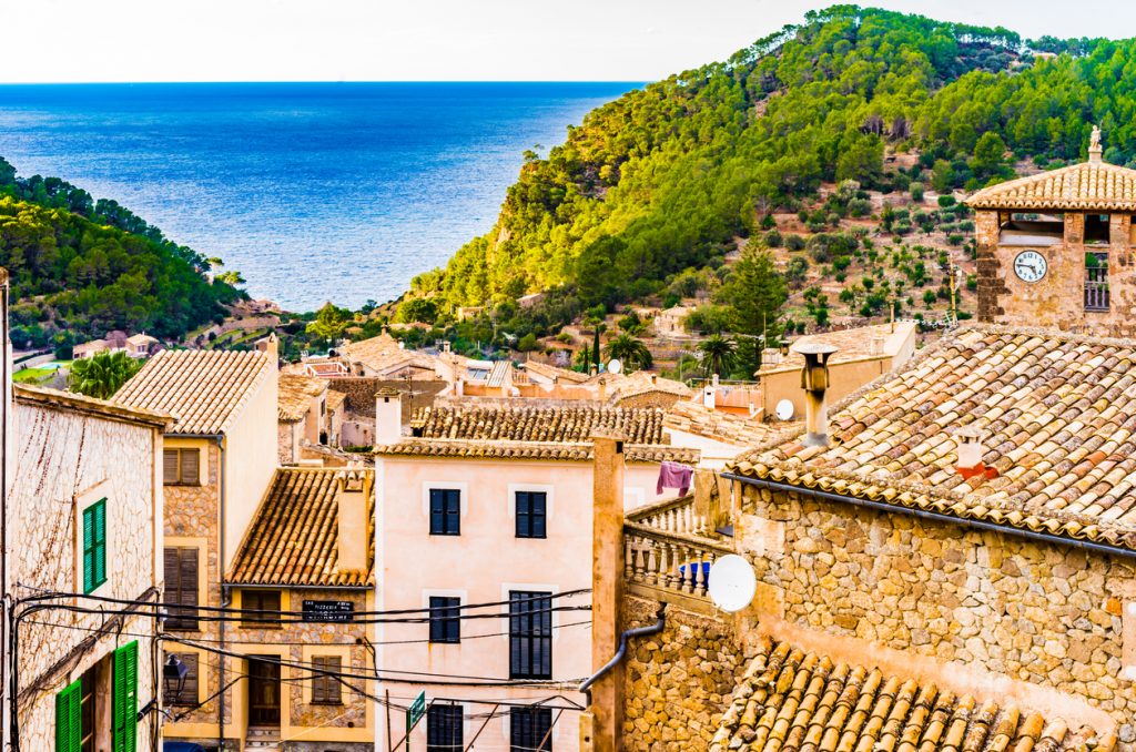 Dächer von Estellencs mit Uhrturm, grünen Hügeln und Meer am Talende.