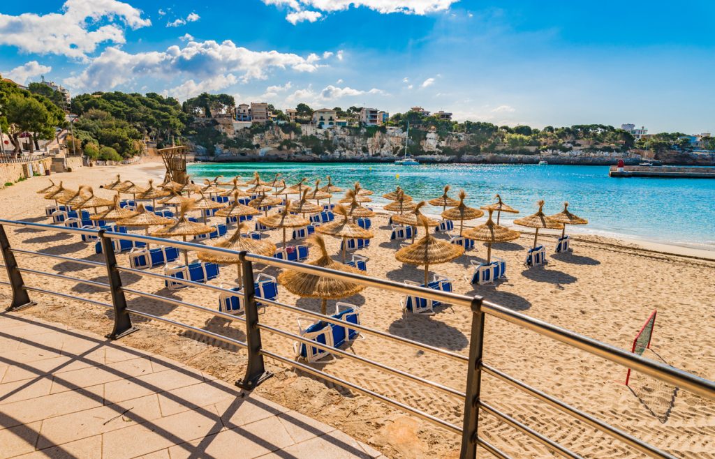 Blick über eine Promenadenbrüstung auf den Sandstrand von Porto Cristo mit Strohschirmen, blauen Liegen und türkisfarbenem, ruhigem Wasser.