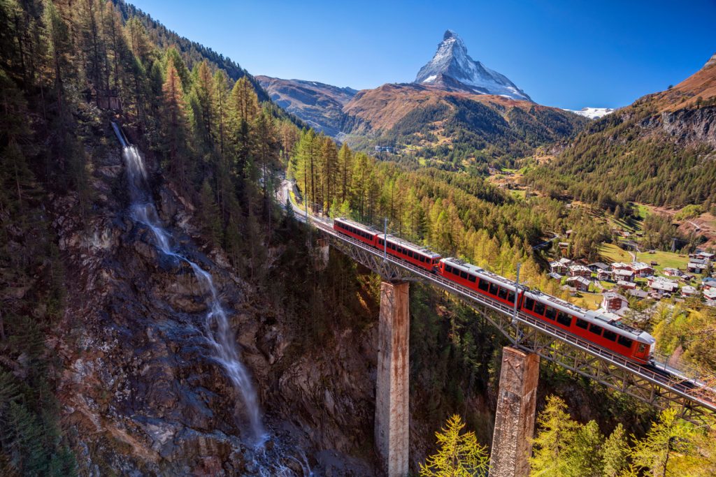 Roter Zug fährt über eine hohe Brücke mit Wasserfall und dem Matterhorn im Hintergrund.
