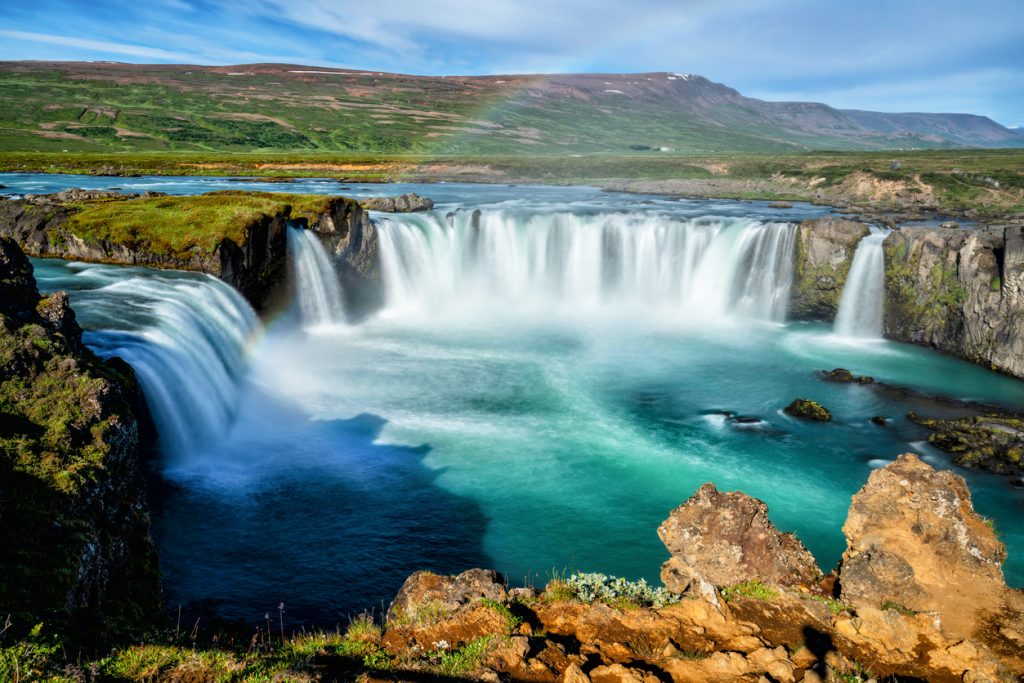 Breiter Wasserfall mit türkisfarbenem Becken und zartem Regenbogen.