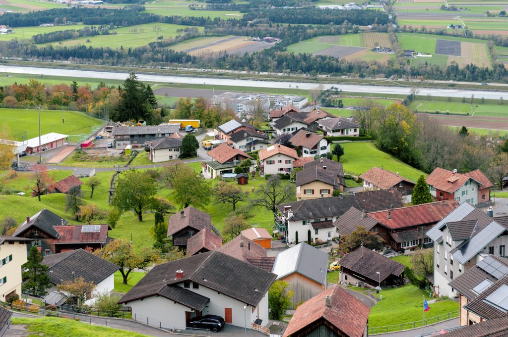 Hügelige Siedlung mit traditionellen Häusern, Wiesen und dem Rhein im Hintergrund.