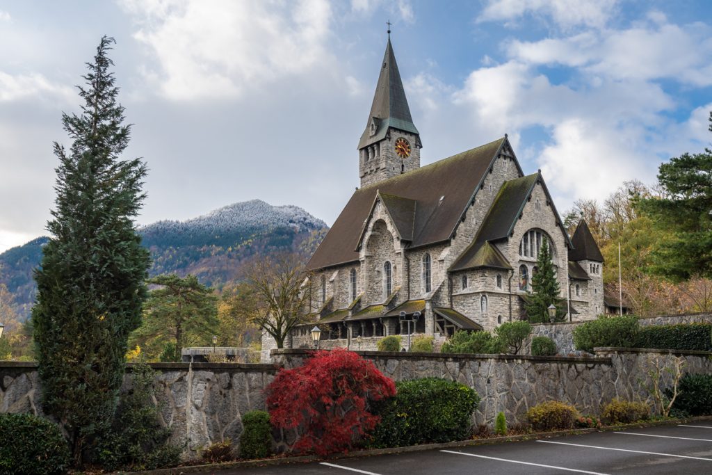 Steinerne Kirche mit hohem Turm in Balzers, davor Gartenmauer und Bäume, hinten leicht verschneite Berge.