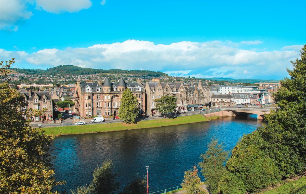 Uferpromenade mit viktorianischen Häusern und Brücke in Inverness unter blauem Himmel.