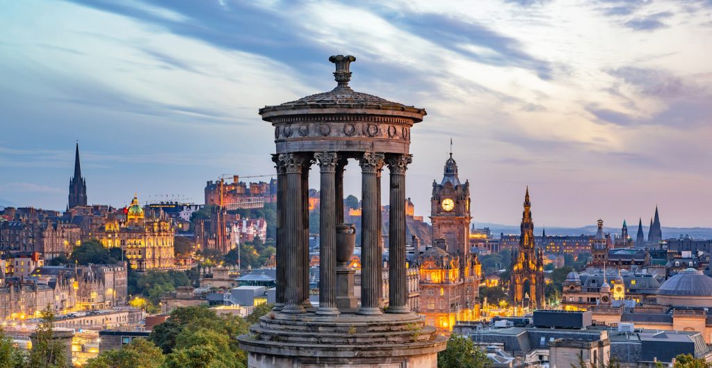 Rundes Monument im Vordergrund und Edinburghs Skyline mit Castle, Balmoral-Uhrturm und Scott Monument in der Dämmerung.